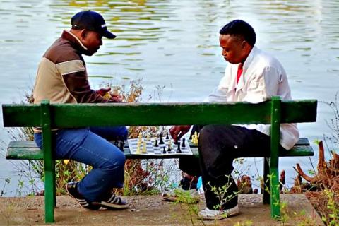 Two men are playing chess on a bench.. The Thai for "Two men are playing chess on a bench." is "ผู้ชายสองคนกำลังเล่นหมากรุกบนม้านั่ง".