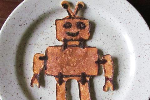 A robot-shaped pancake on a white plate. The Thai for "a robot-shaped pancake on a white plate" is "แพนเค็กรูปหุ่นยนต์บนจานสีขาว".