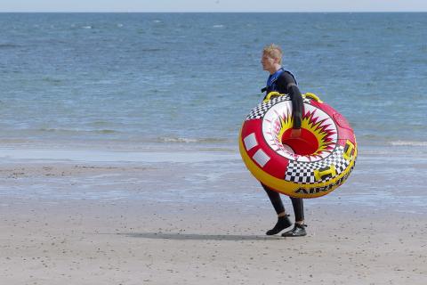 A man is carrying a rubber ring at the beach. The Thai for "a man is carrying a rubber ring at the beach" is "ผู้ชายถือห่วงยางที่ชายหาด".