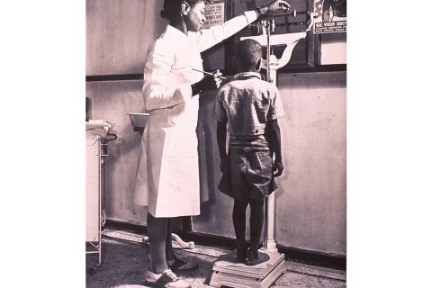 A nurse weighing a boy. The Thai for "a nurse weighing a boy" is "พยาบาลชั่งน้ำหนักเด็กผู้ชาย".