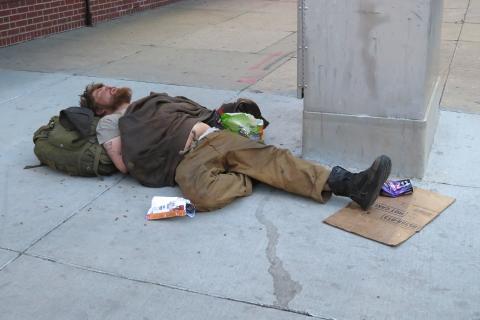 A homeless man sleeping on the street. The Thai for "a homeless man sleeping on the street" is "คนไร้บ้านนอนหลับบนถนน".