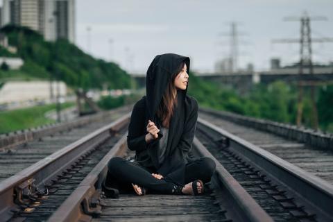 A woman in a hoodie sitting on the railroad. The Thai for "a woman in a hoodie sitting on the railroad" is "ผู้หญิงในเสื้อคลุมมีหมวกนั่งบนทางรถไฟ".