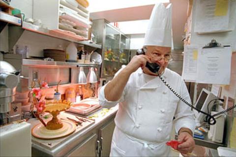A chef speaking on a telephone in the kitchen. The Thai for "a chef speaking on a telephone in the kitchen" is "พ่อครัวพูดโทรศัพท์ในห้องครัว".